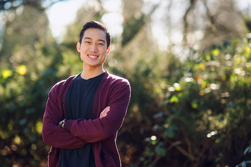 Casey Chung stands with his arms crossed in front of a forested background
