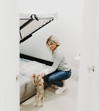 Jane Stoller organizes a bedroom by neatly storing items in a compartment under a bed while her dog watches