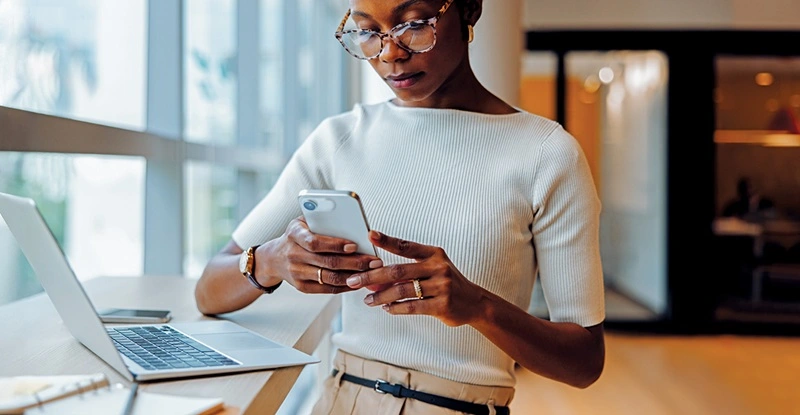Businesswoman reading her smartphone while working on her laptop at a desk