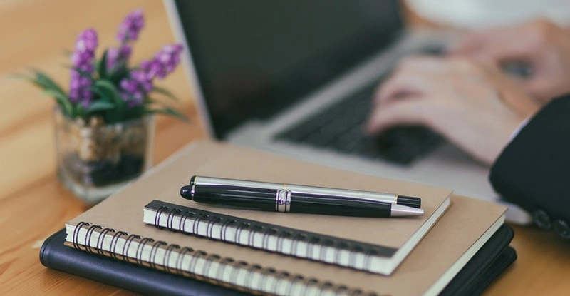 Person working at a laptop on a desk with a stack of notebooks and pens next to them