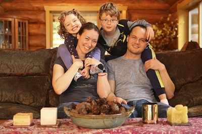 Jodi Moss and her partner sit on their couch with each one holding one of their children on their shoulders. They are all smiling at the camera